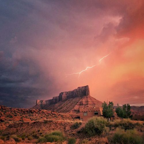 A dramatic desert landscape with a rocky mesa, vibrant sunset, storm clouds, and a lightning strike, set behind a small house and sparse vegetation.
