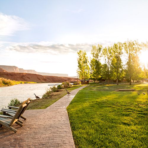 A scenic riverside view with chairs on a paved path, trees, mountains, clear sky, and sunlight shining through the trees in a serene environment.