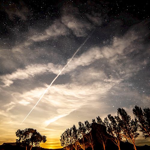 A night sky with starry clouds and a bright streak, possibly a meteor or airplane trail, above a silhouette of trees and distant hills.