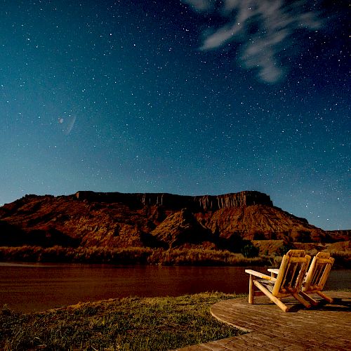 A pair of wooden chairs sits by a river under a starry night sky, with a mountainous landscape in the background.