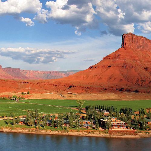 The image shows a panoramic view of a river winding through a scenic valley with green fields and a settlement, surrounded by red sandstone mesas and buttes.
