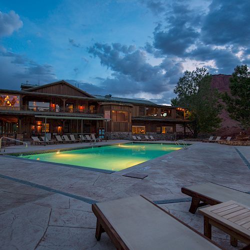 A tranquil evening view of a resort with an illuminated swimming pool, lounge chairs, and a rustic lodge in the background under a cloudy sky.