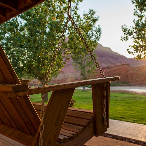 A wooden porch swing hangs with a scenic view of a river, trees, and mountains at sunset. The sun casts a warm glow over the tranquil setting.