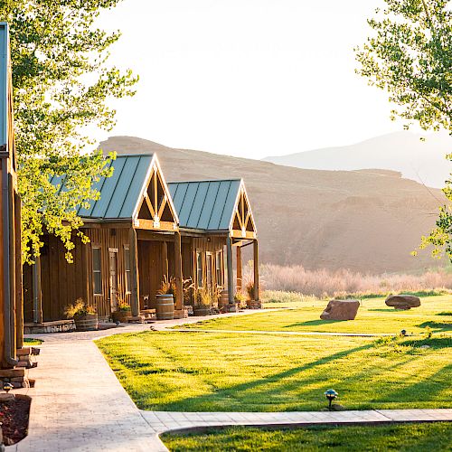 This image shows a scenic outdoor setting with wooden cabins, a lush green lawn, trees, and distant hills, bathed in warm sunlight.