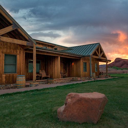 A rustic wooden house with a porch is set against a dramatic sunset and surrounded by a grassy yard with a large red rock in the foreground.