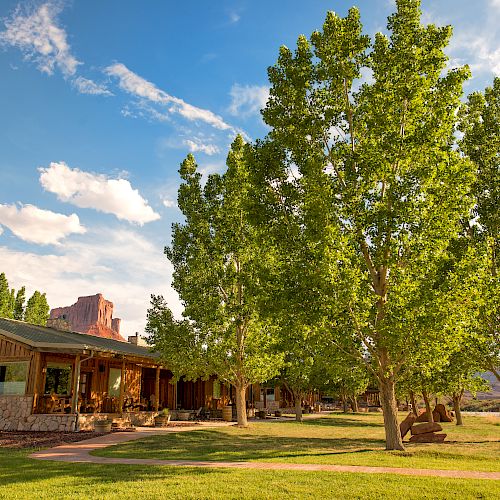 A quaint wooden building with a green roof is surrounded by tall trees and lush grass under a bright, partly cloudy sky.
