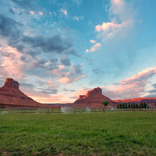 A scenic view of two large mesas under a colorful sky with a grassy field and some trees in the foreground, partly cloudy sky.