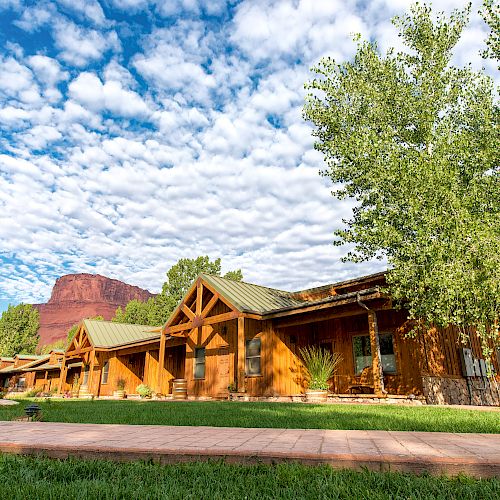 The image shows a rustic wooden lodge set against a stunning backdrop of red rock cliffs under a partly cloudy sky, with greenery around.