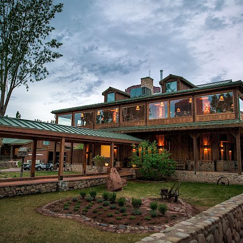 A rustic two-story building with large windows, surrounded by trees and landscaping, under an overcast sky, featuring a stone fence and pathway.