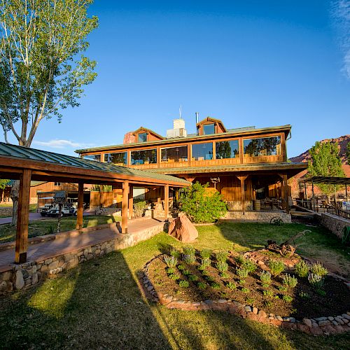 A rustic building with large windows and a covered walkway is surrounded by greenery and scenic mountains under a clear blue sky.