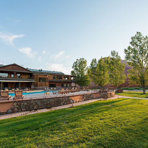 An outdoor scene featuring a large, wooden building, a swimming pool with lounge chairs, and a grassy lawn with trees under a clear sky.