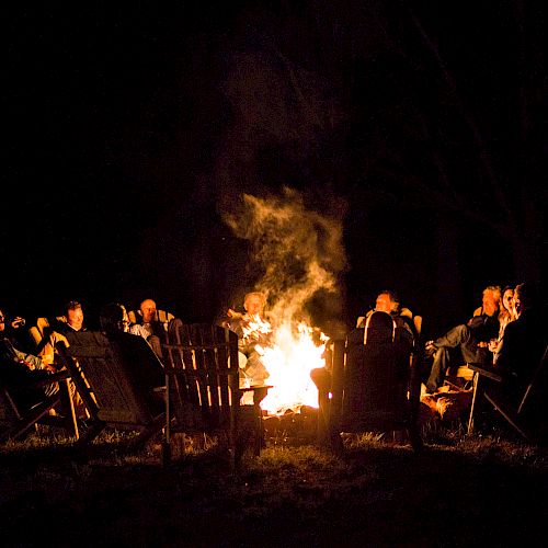 A group of people sitting around a campfire at night, enjoying each other's company in a relaxed outdoor setting, with chairs arranged in a circle.