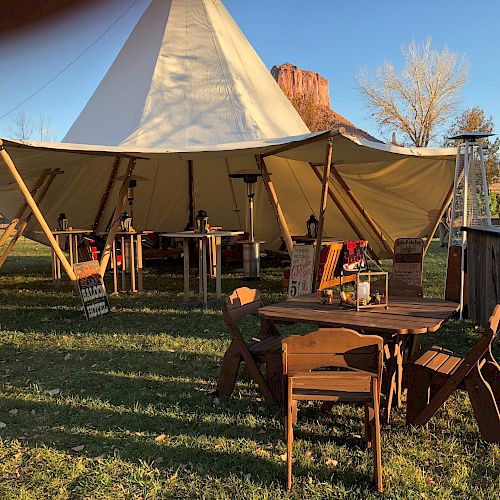 An outdoor setup features a large tent with a canopy, surrounded by wooden furniture including tables and chairs, in a grassy area with trees.