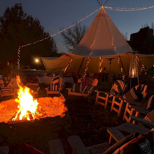 A cozy nighttime campsite with a large tent, a fire pit surrounded by chairs, and string lights illuminating the scene.