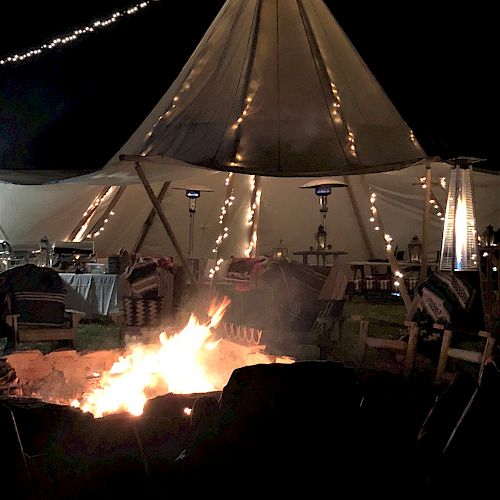 A nighttime scene with a large glowing tipi decorated with string lights and a fire pit in front, surrounded by chairs.