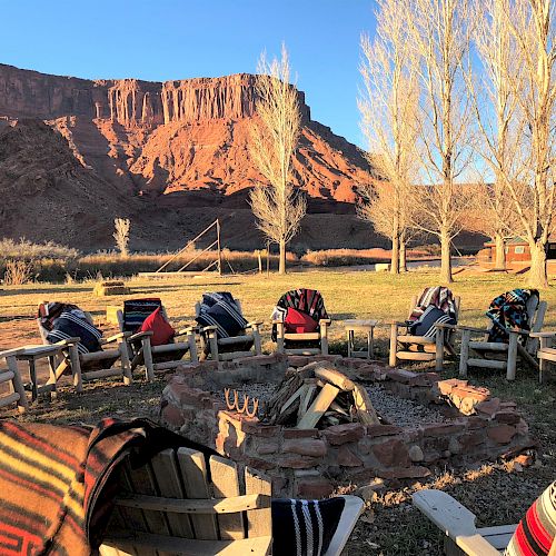 A circular arrangement of wooden chairs with blankets draped over them around an outdoor fire pit, set against a backdrop of rocky cliffs.