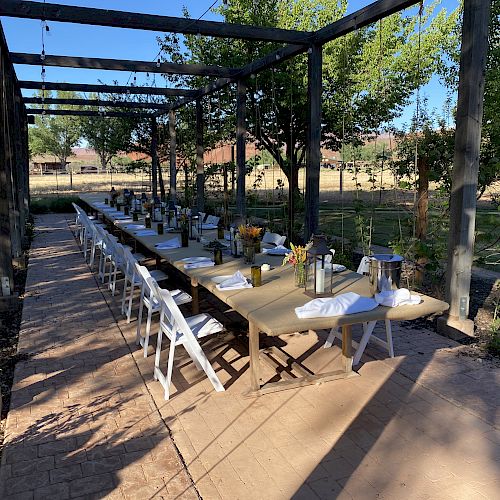 The image shows an outdoor dining area with a long table set for a meal, surrounded by chairs under a pergola, in a garden setting.