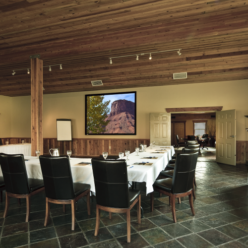 A conference room with a long table and black chairs, a large screen displaying a landscape image, and a refreshment table by the window.