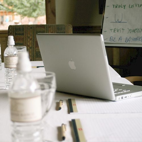 A conference table with bottled water, documents, and pens is seen. A laptop is open, and a whiteboard with text is in the background.