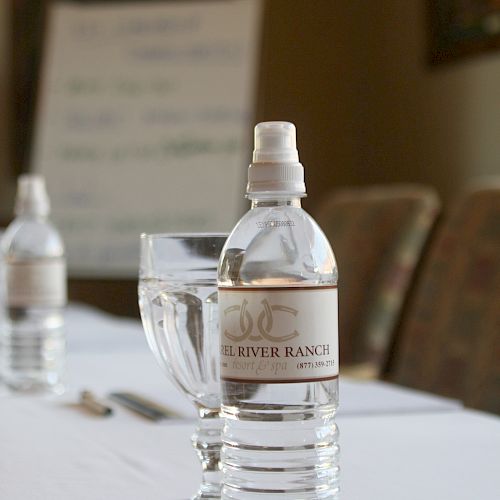 The image shows a bottle of water, a glass of water, and a blurred whiteboard in a meeting room setting, placed on a white tablecloth.