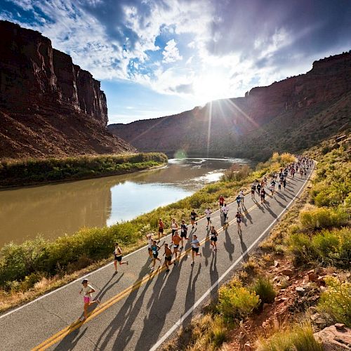 A group of runners is participating in a race on a scenic road next to a river, with sunlight streaming through the clouds and a canyon in the background.