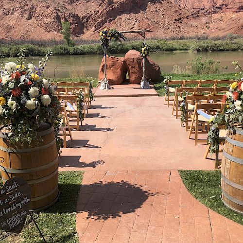 An outdoor wedding setup with wooden chairs, flower arrangements on barrels, and an arch with flowers by a river, set against a desert backdrop.