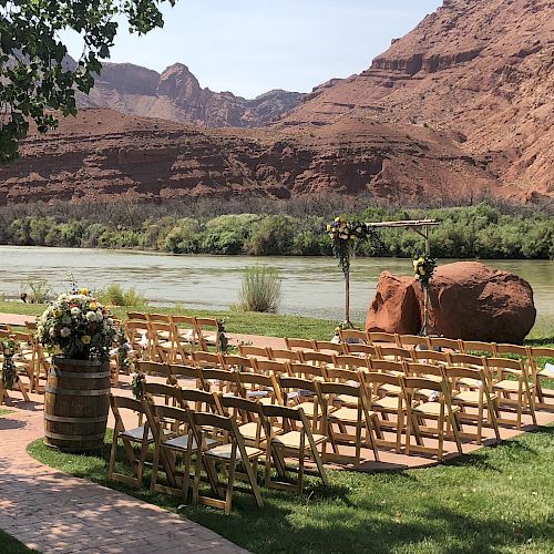 An outdoor wedding setup beside a river, with rows of wooden chairs, floral arrangements, and a wooden archway under clear skies and mountains.
