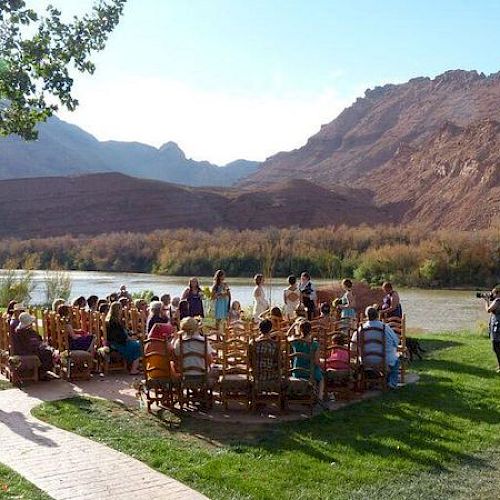 A group of people is gathered outdoors near a river, with mountains in the background, likely attending a ceremony or event.