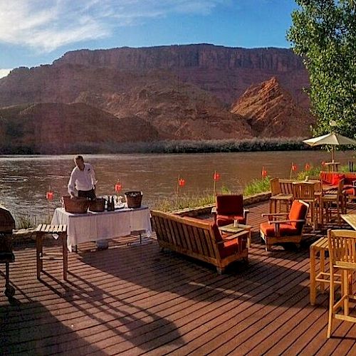 A scenic riverside deck with outdoor seating, tables, umbrellas, and a person preparing food near a grill and serving station. Rocky mountains in the background.