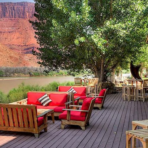 An outdoor deck with red cushioned seating, wooden tables, and chairs overlooking a scenic river and canyon landscape.