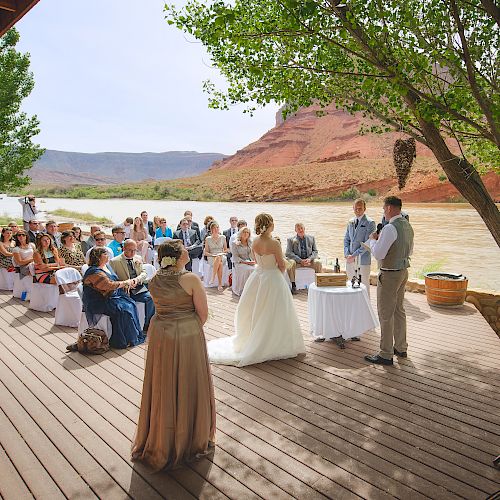 A wedding ceremony is taking place outdoors by a river, with guests seated, the bride and groom standing, and a scenic background of red mountains.