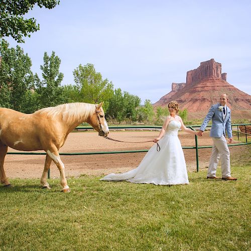 A bride and groom, dressed for their wedding, guide a horse on a grassy field with red rock formations in the background.