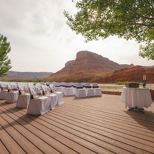 An outdoor wedding setup by a river with chairs covered in white fabric, a round table with candles, and a scenic mountain in the background.