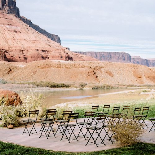 A scenic outdoor setting with folding chairs arranged in rows on a patio, overlooking a river and surrounded by rugged, rocky terrain and cliffs.