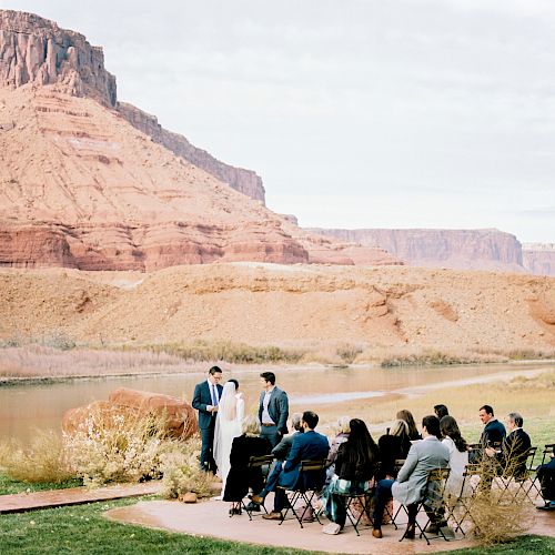 A small wedding ceremony is taking place outdoors with red cliffs and a river in the background. Guests are seated, and the couple stands by an officiant.