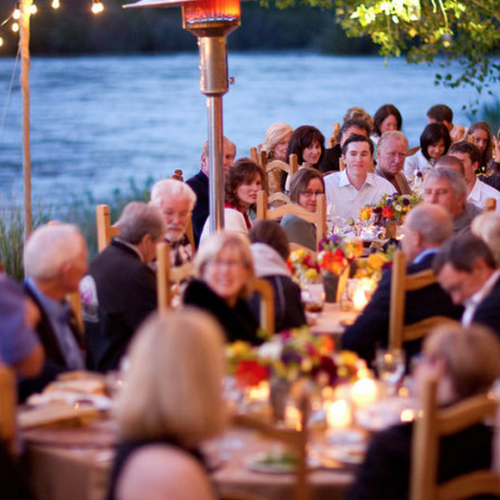 People are seated at an outdoor event with tables decorated with flowers and lights, listening to someone speaking. It looks like a formal gathering.