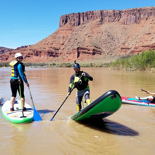 Four people are paddleboarding and kayaking on a river surrounded by red rock cliffs and a clear blue sky in the background.