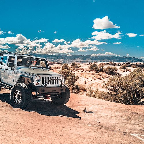 A jeep is parked on a rocky terrain under a bright blue sky with scattered clouds, and a vast desert landscape in the background.