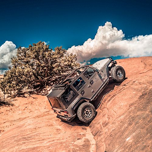 A rugged off-road vehicle climbing a steep, rocky terrain under a clear blue sky with scattered clouds and surrounded by some bushes and trees.