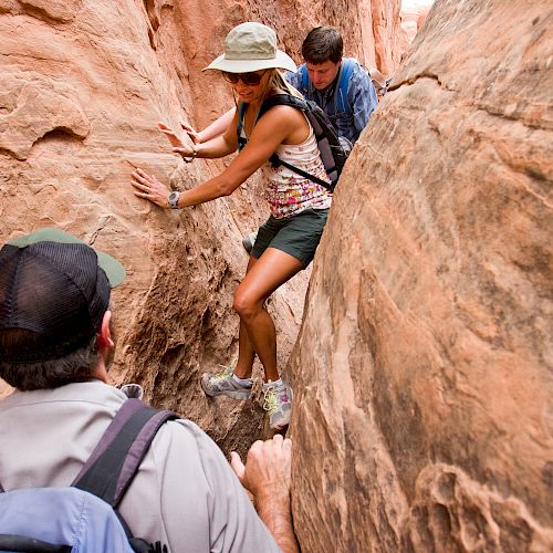 Three people navigate a narrow slot canyon, with one person climbing ahead and two following behind through the tight, rocky passage.