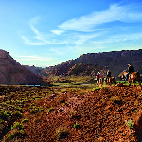 A group of people is horseback riding on a trail through a desert landscape with mountains and a bright, clear sky in the background.
