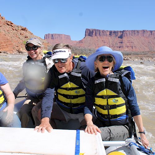 Four people wearing life jackets are seated in a boat, enjoying a river rafting trip with red rock formations and clear blue sky in the background.