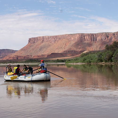 A group of people are on a rafting trip in a calm river, with scenic red rock cliffs and clear skies in the background.