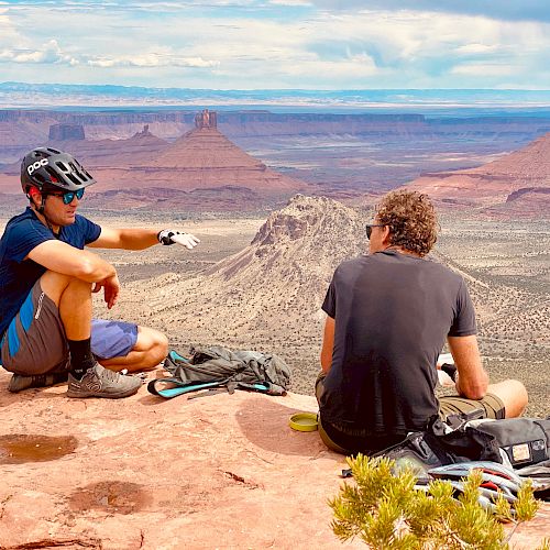 Two people in cycling gear rest on a rocky cliff overlooking a vast desert landscape with mesas and buttes in the distance, under a partly cloudy sky.