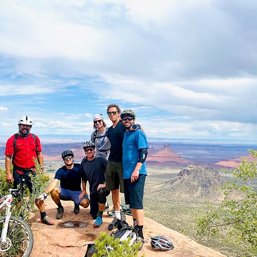 A group of five people in biking gear standing on a scenic overlook with desert mountains in the background, and bicycles in the foreground.