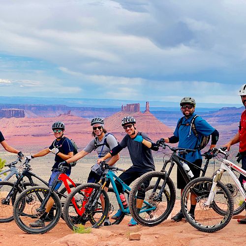 A group of six people in cycling gear stand with their bikes on a red rock landscape, with scenic canyons and mesas in the background under a cloudy sky.
