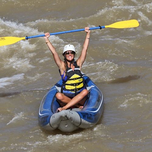 A person in a helmet and life jacket is sitting in a kayak, holding a paddle overhead while navigating rapids on a river.