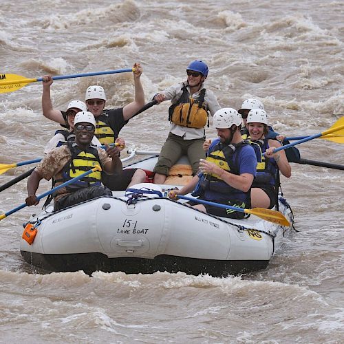 A group of people in life vests and helmets are white-water rafting on a river, holding yellow paddles and appearing excited.