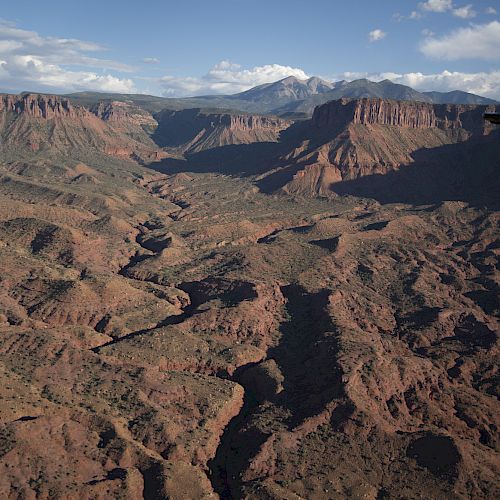 An aerial view of a vast, rocky canyon landscape under a clear blue sky, with distant mountains and scattered clouds in the background.