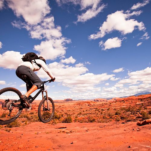 A cyclist rides a mountain bike on a rugged, red rock desert terrain under a vibrant, partly cloudy blue sky.